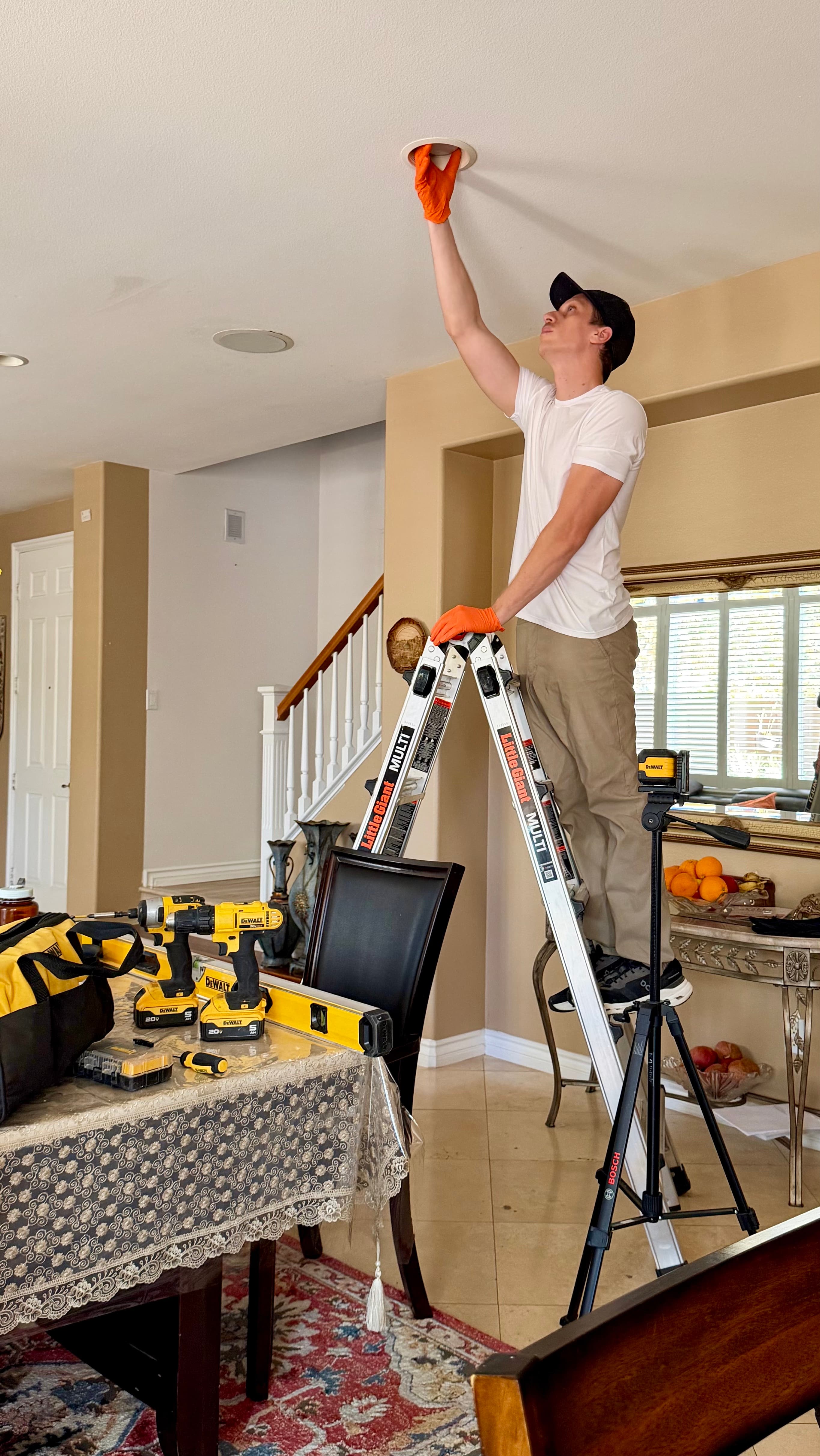 Man on a ladder installs a recessed ceiling light with tools on a dining table.