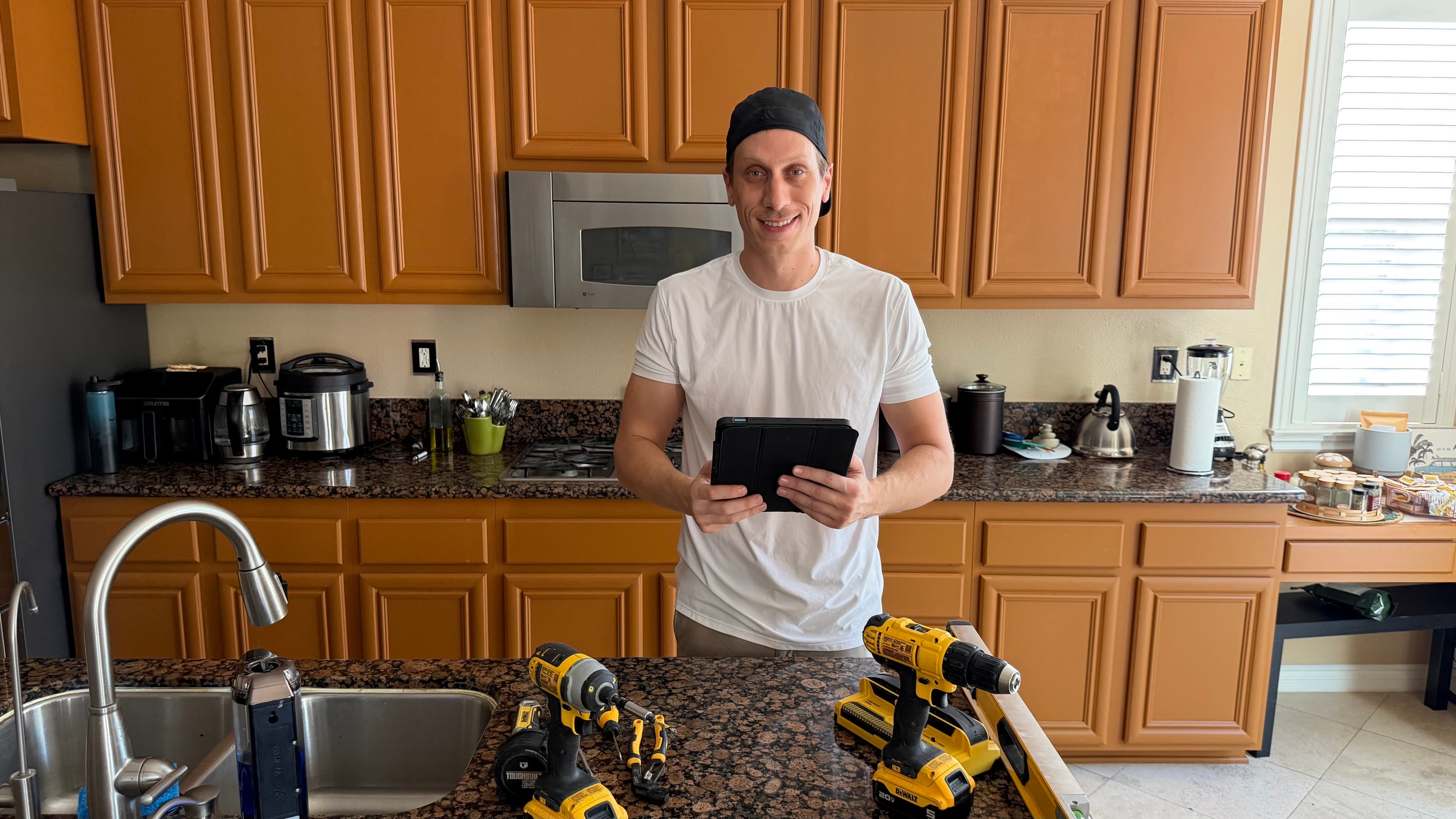 Smiling man in a kitchen holding a tablet with power tools on the granite counter.